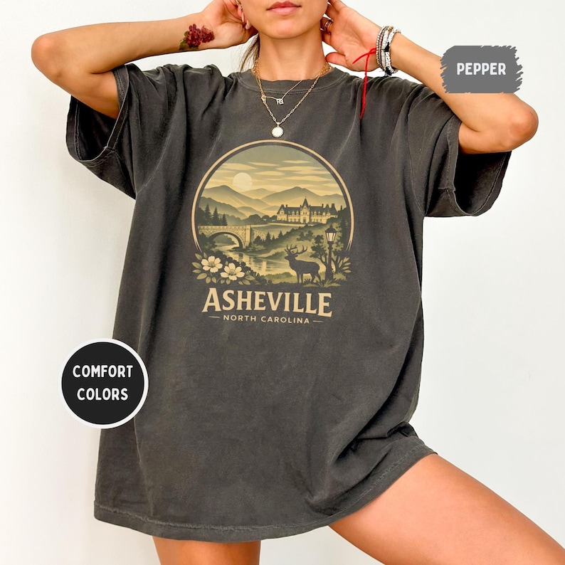 May include: Dark grey t-shirt with a circular graphic featuring a bridge, mountains, and a deer, with the text "Asheville North Carolina". The shirt is labeled "Comfort Colors" and "Pepper".