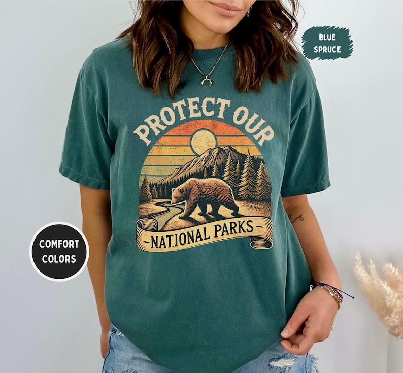 May include: A teal blue t-shirt with a vintage-style graphic of a bear in a forest scene. The graphic says "Protect Our National Parks".
