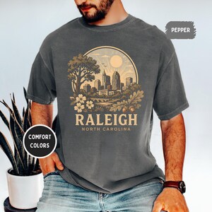 May include: Dark gray t-shirt with a circular graphic featuring a cityscape, tree, and flowers. The word "RALEIGH" is printed below the graphic, with "NORTH CAROLINA" underneath. The shirt has short sleeves and a crew neck.
