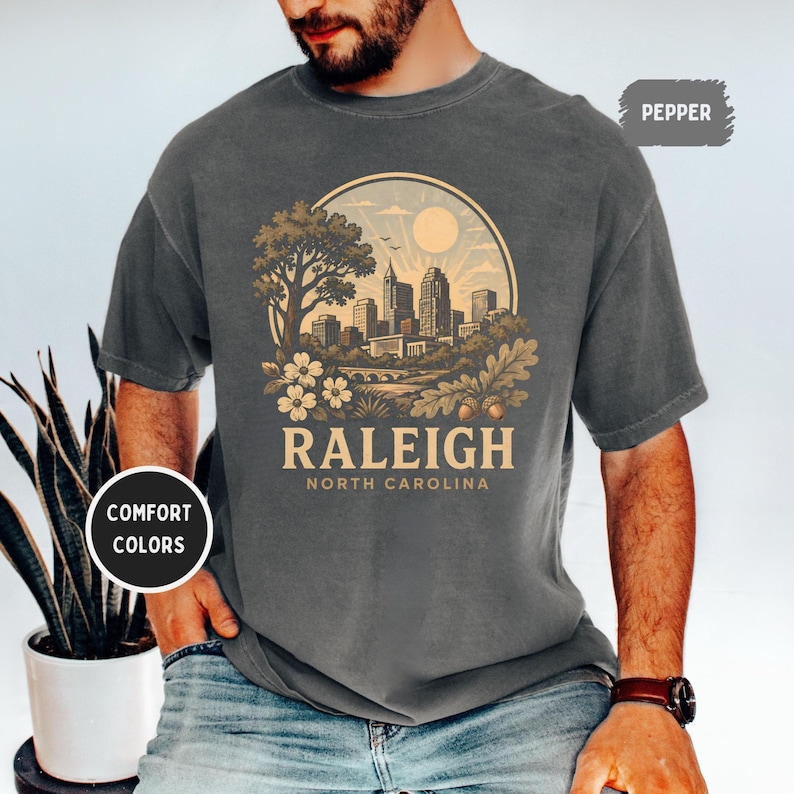 May include: Dark gray t-shirt with a circular graphic featuring a cityscape, tree, and flowers. The word "RALEIGH" is printed below the graphic, with "NORTH CAROLINA" underneath. The shirt has short sleeves and a crew neck.