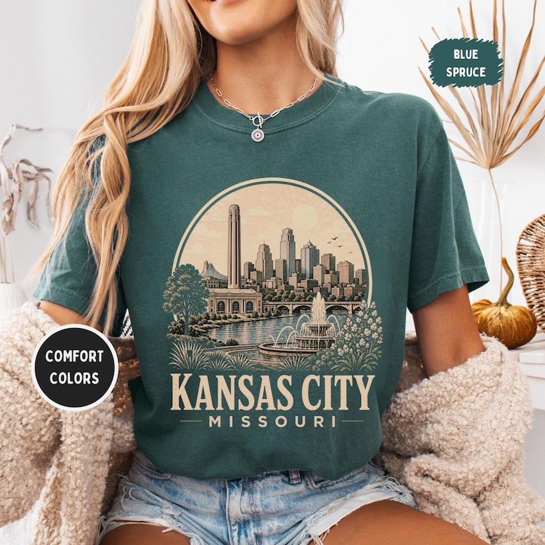 May include: Blue spruce t-shirt with a graphic of the Kansas City skyline, a fountain, and the text "Kansas City Missouri". The shirt has a round neck and short sleeves. The words "Comfort Colors" are printed on a white circle.