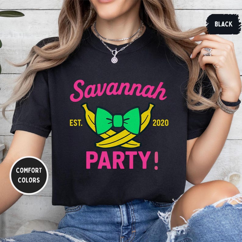 May include: Black t-shirt with the text "Savannah PARTY!" in pink. The design includes a green bow tie and two yellow bananas. The text "EST. 2020" is also present. The shirt is labeled "COMFORT COLORS".