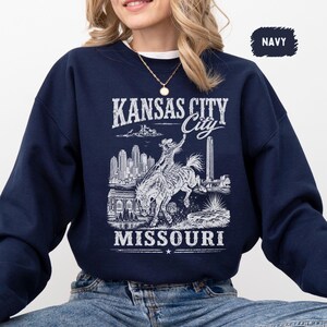 May include: Navy blue sweatshirt featuring a white graphic of Kansas City, Missouri. The design includes the city skyline, a cowboy riding a horse, and the text "KANSAS CITY" and "MISSOURI". The word "NAVY" is in a small box in the upper right corner.