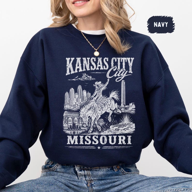 May include: Navy blue sweatshirt featuring a white graphic of Kansas City, Missouri. The design includes the city skyline, a cowboy riding a horse, and the text "KANSAS CITY" and "MISSOURI". The word "NAVY" is in a small box in the upper right corner.