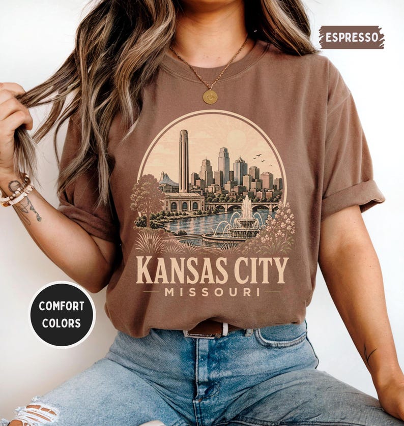 May include: Espresso-colored t-shirt featuring a circular graphic of the Kansas City skyline, a bridge, and a fountain. The text "KANSAS CITY MISSOURI" is printed below the graphic. The shirt is part of the Comfort Colors collection.