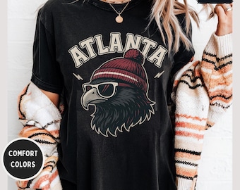 May include: Black t-shirt with a graphic of an eagle wearing sunglasses and a maroon beanie. The word "ATLANTA" is arched above the eagle in a vintage font. The shirt has a distressed look and the words "COMFORT COLORS" are in a circle.