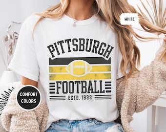 May include: White t-shirt with a distressed graphic design. The design features the text "PITTSBURGH FOOTBALL" above a football graphic and the text "ESTD. 1933". The shirt has the words "COMFORT COLORS" and "WHITE" printed on it.