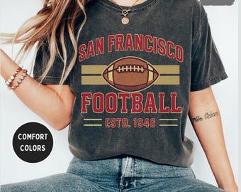 May include: Dark grey t-shirt featuring the text "SAN FRANCISCO FOOTBALL" in red above a brown football graphic. The word "FOOTBALL" is in red below the graphic, with "ESTD. 1946" beneath. The shirt is paired with blue jeans.