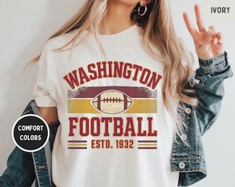 Op de afbeelding: Ivoor t-shirt met een distressed design. Het heeft "WASHINGTON FOOTBALL" in bordeauxrood en goud, met een voetbalafbeelding boven "FOOTBALL ESTD. 1932". Het shirt maakt deel uit van de Comfort Colors collectie.