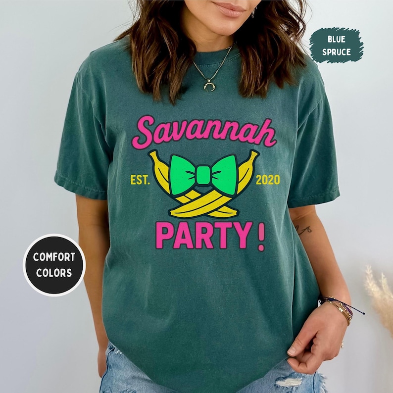 May include: Blue spruce t-shirt with the text "Savannah PARTY!" in pink and a green bow tie over yellow bananas. The text "EST. 2020" is also included. The shirt is a comfort colors brand.
