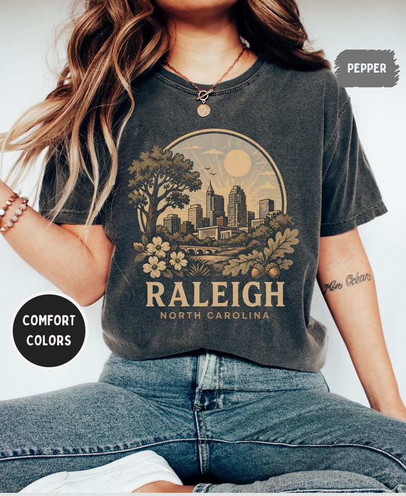 May include: Dark gray t-shirt featuring a circular graphic of the Raleigh, North Carolina skyline, with a tree, flowers, and the text "RALEIGH NORTH CAROLINA." The shirt has short sleeves and a crew neck. The words "COMFORT COLORS" and "PEPPER" are also visible.