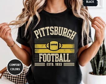 May include: Black t-shirt featuring "Pittsburgh Football" text and a football graphic in yellow and black. The text "ESTD. 1933" is below the graphic. The shirt is a comfort colour.