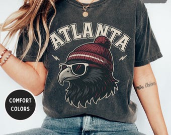 May include: Dark grey t-shirt featuring the word "ATLANTA" in white block letters above a graphic of a hawk wearing sunglasses and a maroon beanie. The shirt has the words "COMFORT COLORS" in a white circle.