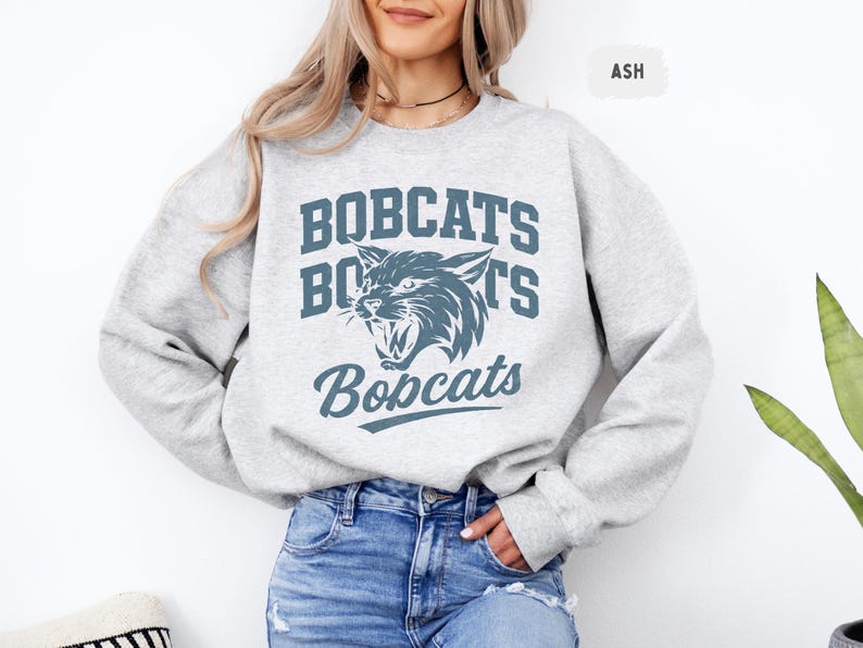 May include: Ash-colored sweatshirt with the word "BOBCATS" in a blue, collegiate-style font. A snarling wildcat graphic is centered below the word. The word "Bobcats" is written in a script font below the graphic.