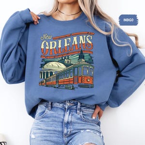 May include: Indigo blue sweatshirt with a graphic print of a streetcar in New Orleans. The text "New Orleans" is printed in white with a red outline.