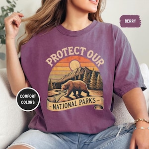 May include: A purple t-shirt with a vintage-style graphic of a bear in a forest with the text "Protect Our National Parks".