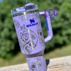 May include: A purple stainless steel tumbler with a black and white skull and floral design. The tumbler has a handle and a straw lid. The Stanley logo is visible on the side of the tumbler.