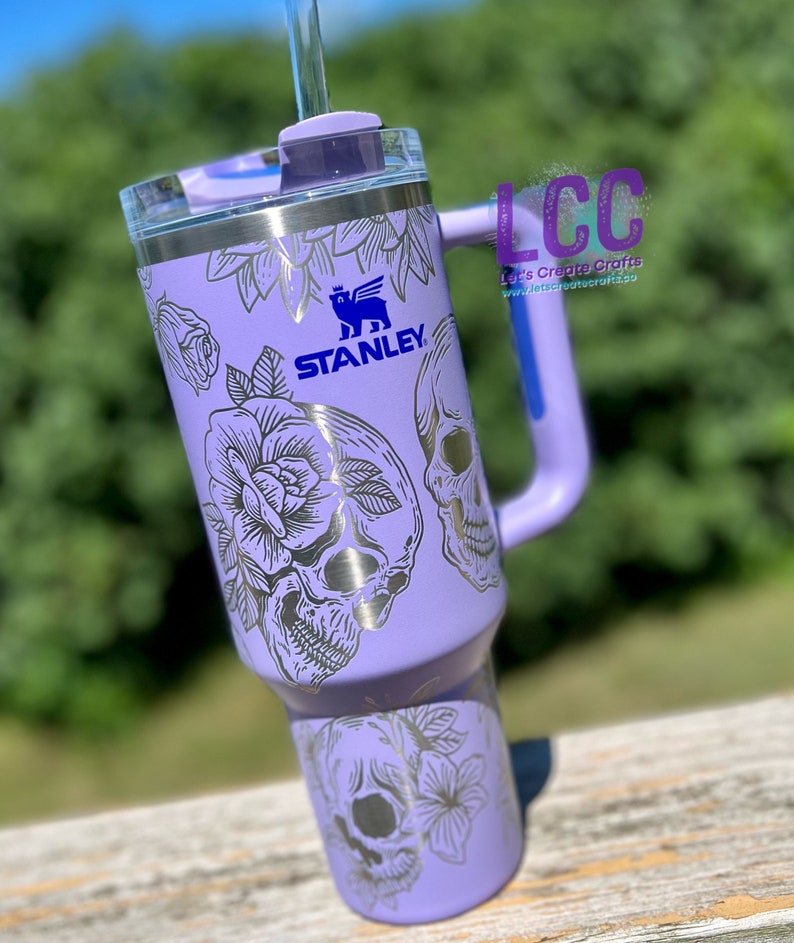 May include: A purple stainless steel tumbler with a black and white skull and floral design. The tumbler has a handle and a straw lid. The Stanley logo is visible on the side of the tumbler.