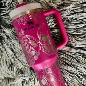 May include: A pink stainless steel travel mug with a black Stanley logo and a silver skull and floral design. The mug has a lid with a straw.
