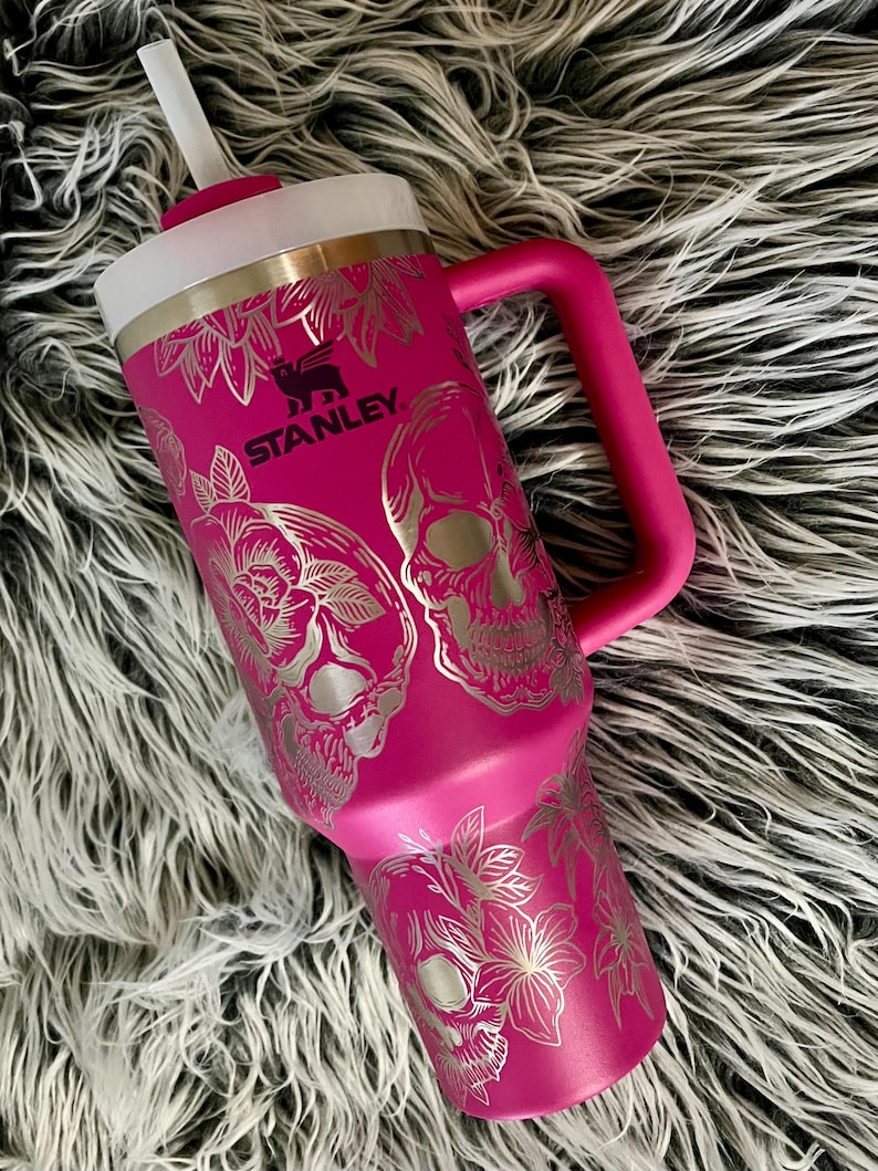 May include: A pink stainless steel travel mug with a black Stanley logo and a silver skull and floral design. The mug has a lid with a straw.