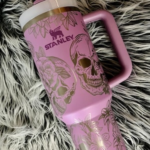 May include: A purple stainless steel travel mug with a handle. The mug has a floral and skull design with the word "STANLEY" printed on the side.