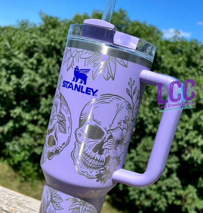 May include: A purple stainless steel tumbler with a black and white skull and floral design. The tumbler has a handle and a lid with a straw. The tumbler is branded with the Stanley logo.