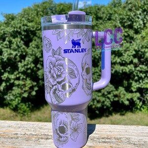 May include: A purple stainless steel tumbler with a black and white skull and floral design. The tumbler has a lid with a straw hole and a handle. The tumbler is branded with the Stanley logo.