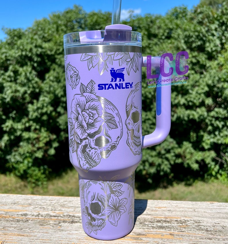 May include: A purple stainless steel tumbler with a black and white skull and floral design. The tumbler has a lid with a straw hole and a handle. The tumbler is branded with the Stanley logo.