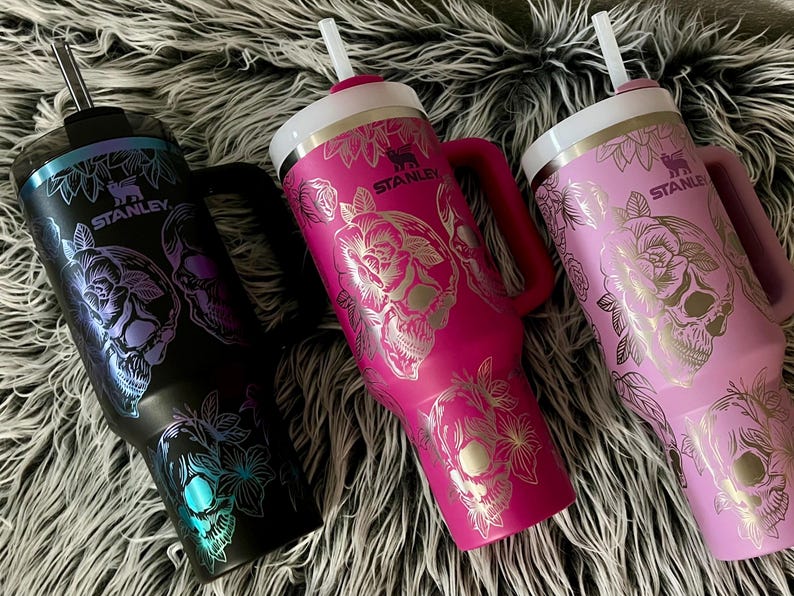 May include: Three stainless steel tumblers with a skull and floral design. The tumblers are in black, pink, and purple. Each tumbler has a straw and a lid. The tumblers have the word "STANLEY" printed on them.