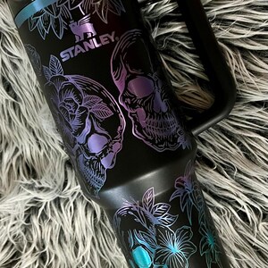 May include: Black stainless steel travel mug with a black handle. The mug has a floral and skull design with iridescent purple and blue accents. The mug has the word "STANLEY" printed on it.