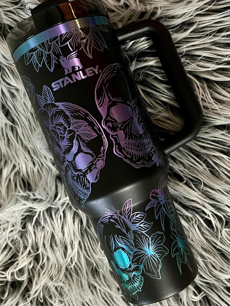 May include: Black stainless steel travel mug with a black handle. The mug has a floral and skull design with iridescent purple and blue accents. The mug has the word "STANLEY" printed on it.
