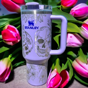May include: A purple stainless steel travel mug with a white skull and floral design. The mug has a black handle and the word "STANLEY" is printed on the side.