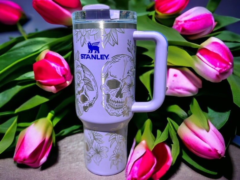 May include: A purple stainless steel travel mug with a white skull and floral design. The mug has a black handle and the word "STANLEY" is printed on the side.