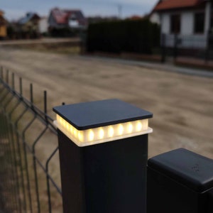 Könnte beinhalten: Ein schwarzer Zaunpfosten mit einer quadratischen Oberseite, auf der sich eine Reihe kleiner weißer LED-Leuchten befinden. Die Leuchten sind beleuchtet und strahlen ein warmes Licht aus.