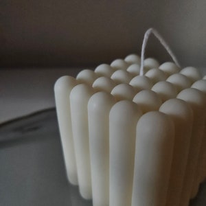 Könnte beinhalten: Eine weiße quadratische Kerze mit einem Docht. Die Kerze besteht aus vielen kleinen, zylindrischen Formen.