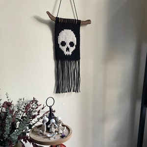 May include: A black and white skull macrame wall hanging with a wooden dowel at the top. The hanging is decorated with black and white fringe. A two-tiered wooden tray with a black metal stand holds a variety of Halloween decorations, including a black skull, a white ghost, and small skeleton figurines.