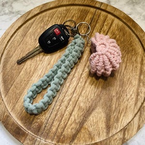May include: A black car key fob attached to a sage green macrame wristlet strap and a small, round, light pink crocheted accessory. The items are connected by a silver-toned metal split ring, showcasing a textured, knotted cord design.