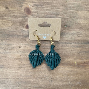 May include: Dark green macrame earrings with gold-colored hooks, displayed on a brown card with the word "Handmade" and a heart. The earrings are leaf-shaped and have a textured appearance. The brand name "MCRMBYAMBER" is printed on the card.