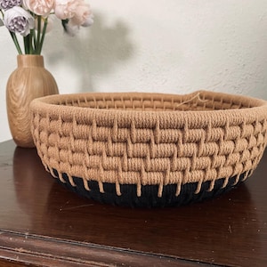May include: A round, woven basket featuring a textured, tan-colored upper section and a solid black base. The basket is constructed with a thick, cord-like material in a repeating grid pattern, creating a sturdy, bowl-shaped container for storage.