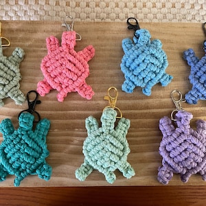 May include: Seven crocheted turtle keychains in various colors, including green, pink, blue, teal, and purple. The keychains are made with a macrame technique and feature a knotted design.