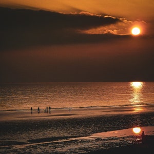 May include: Silhouettes of people walking on a sandy beach at sunset with a large orange sun in the sky.