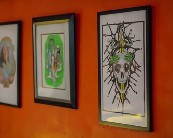 May include: Three framed illustrations on an orange wall. The illustration on the right is a black and white skull with a dagger through it and green leaves around the skull. The illustration in the middle is a green, yellow, and black illustration of a figure with a crown. The illustration on the left is a black and white illustration of a woman with a crown.