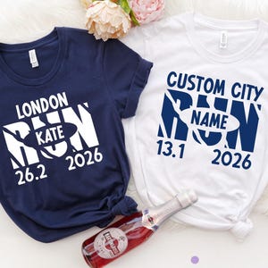 May include: A navy blue t-shirt with white text reading LONDON RUN KATE 26.2 2026 and a white t-shirt with navy blue text reading CUSTOM CITY RUN NAME 13.1 2026, accompanied by a bottle of Martini & Rossi sparkling wine with a red label.