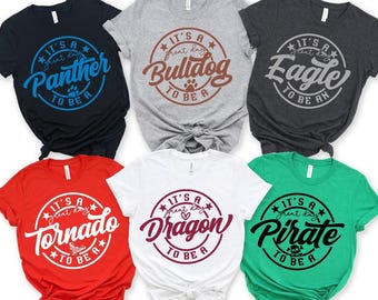 May include: Six short-sleeved t-shirts in black, gray, dark gray, red, white, and green. Each shirt features a circular graphic with the text 'IT'S A GREAT DAY TO BE A' followed by the words Panther, Bulldog, Eagle, Tornado, Dragon, or Pirate.