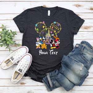 Puede incluir: Camiseta gris oscuro con un gr&aacute;fico colorido de personajes de Disney, un castillo y el a&ntilde;o 2026. El texto "Your Text" est&aacute; debajo. La camiseta se combina con vaqueros azules y zapatillas blancas.