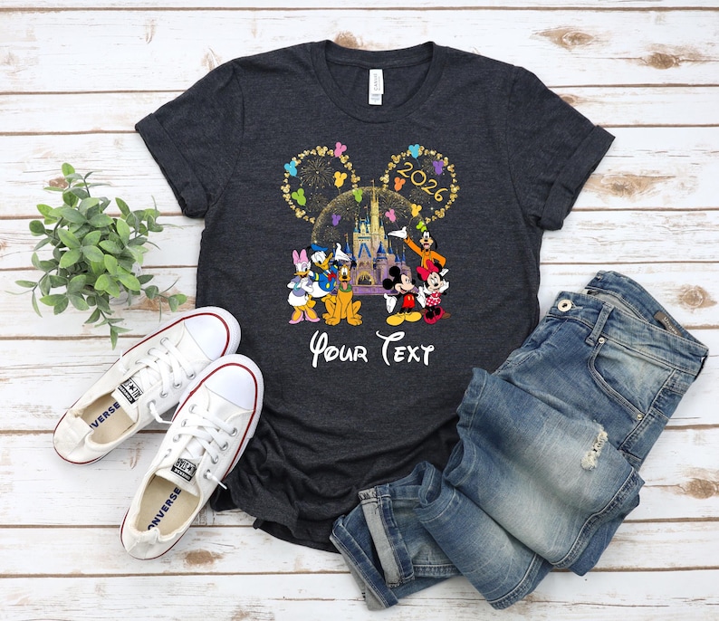 Puede incluir: Camiseta gris oscuro con un gr&aacute;fico colorido de personajes de Disney, un castillo y el a&ntilde;o 2026. El texto "Your Text" est&aacute; debajo. La camiseta se combina con vaqueros azules y zapatillas blancas.