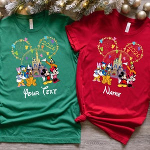 Puede incluir: Dos camisetas, una verde y otra roja, con un dise&ntilde;o de Disney. El dise&ntilde;o incluye orejas de Mickey Mouse, un castillo y varios personajes. La camiseta verde tiene "Your Text", la camiseta roja tiene "Name", y el a&ntilde;o 2026.