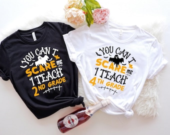 Puede incluir: Una camiseta negra y una camiseta blanca, cada una con un gráfico de fantasma y el texto "YOU CAN'T SCARE ME I TEACH 2ND GRADE" o "YOU CAN'T SCARE ME I TEACH 4TH GRADE" en letras naranjas, blancas y negras con pequeñas siluetas de murciélagos.
