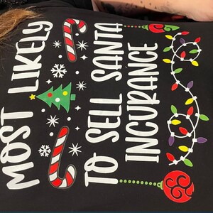 May include: Black t-shirt with white text that reads "Most Likely to Sell Santa Insurance". The text is surrounded by Christmas-themed graphics, including a candy cane, a Christmas tree, snowflakes, and a string of lights.