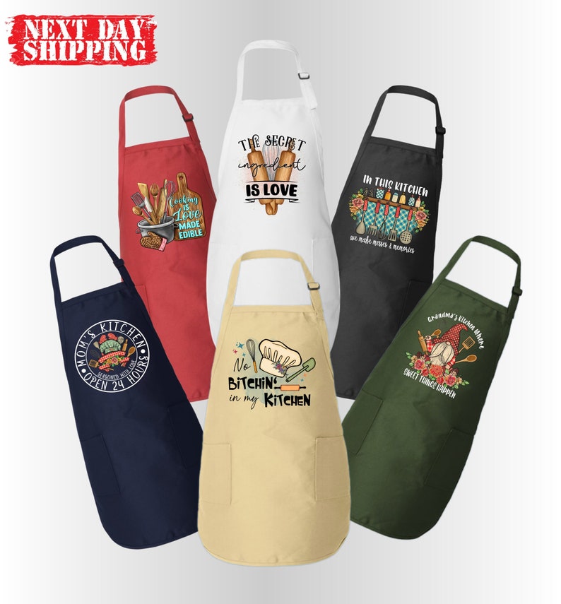 May include: Six different kitchen aprons with various designs and sayings. The aprons are in red, white, black, beige, green, and navy blue. The sayings on the aprons include "The secret ingredient is love", "No bitchin' in my kitchen", "In this kitchen we make memories & miracles", and "Grandma's kitchen shack".
